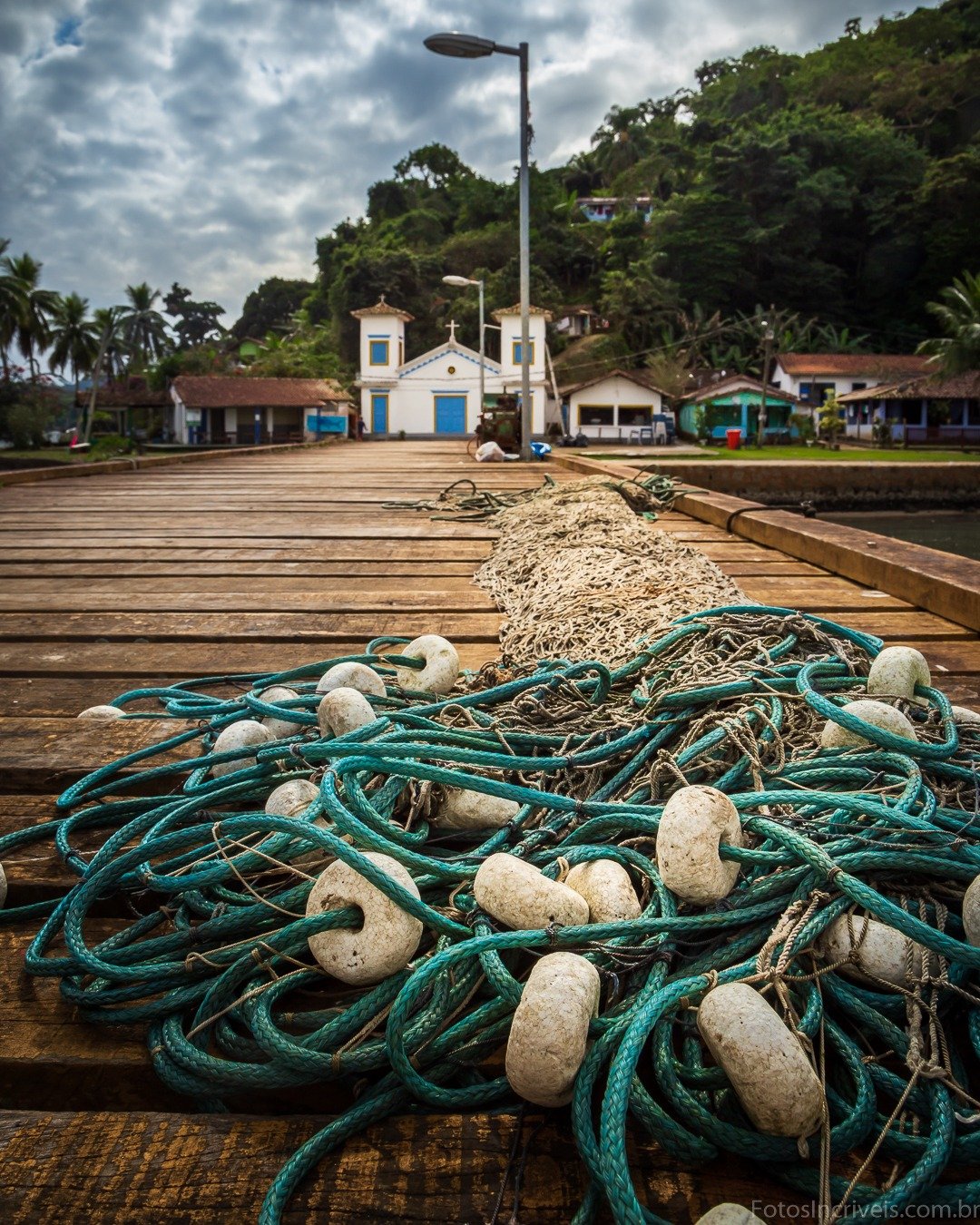Pier de madeira em primeiro plano coberto por rede de pesca verde com boias brancas, tendo ao fundo casario colonial, morro com vegetação densa e céu nublado dramático.