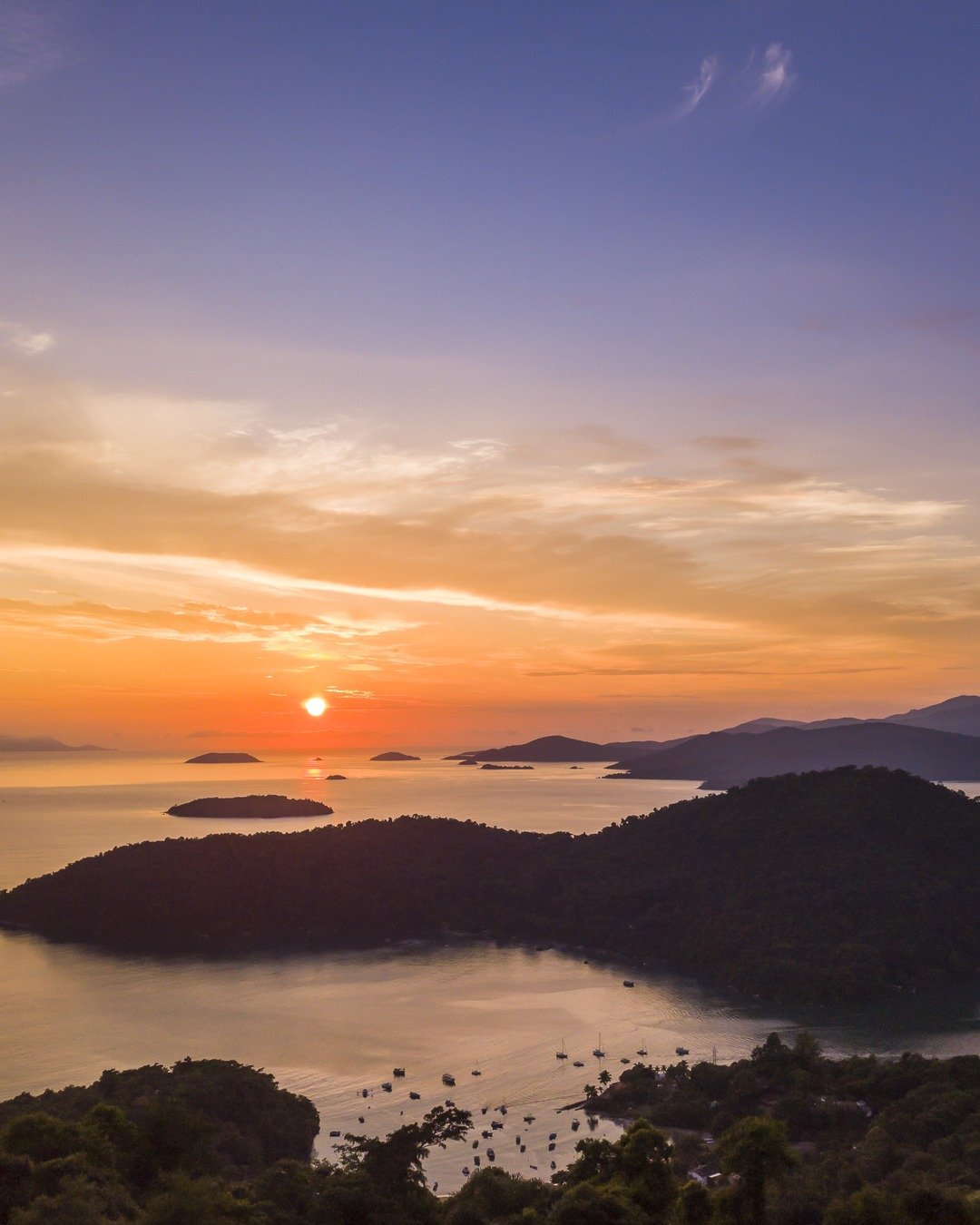 Vista elevada de um amanhecer sobre baía com ilhas e montanhas em silhueta, céu alaranjado refletido na água calma e mata no primeiro plano.