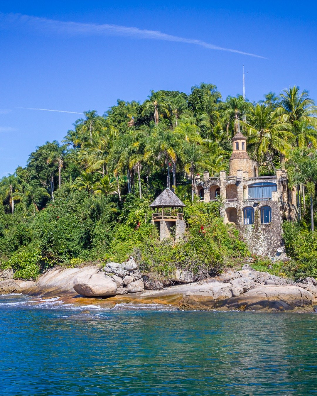 Ruína de construção histórica em pedra sobre ilha rochosa coberta por densa vegetação tropical e palmeiras, cercada por mar azul e céu limpo.