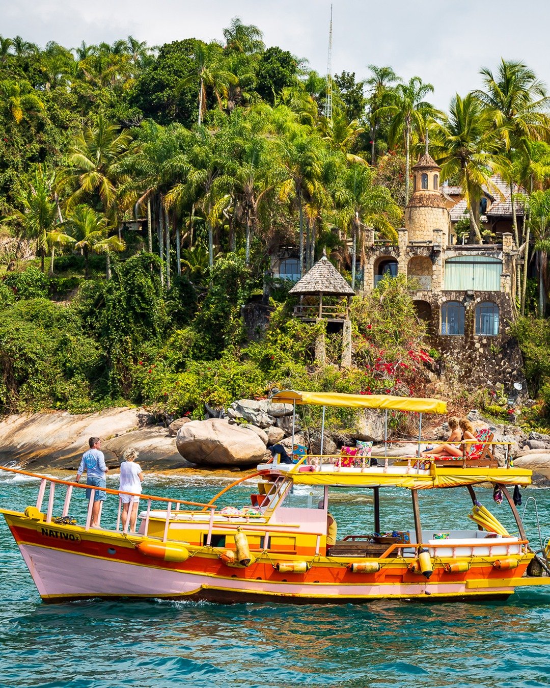Barco amarelo com turistas navegando em água turquesa diante de uma encosta rochosa coberta de palmeiras, com uma construção de estilo castelo entre a vegetação tropical.