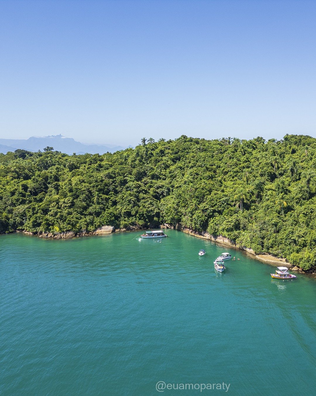 Vista aérea de uma enseada de águas turquesa cercada por morros cobertos de mata atlântica, com pequenas embarcações ancoradas junto à costa e montanhas ao fundo sob céu azul limpo.