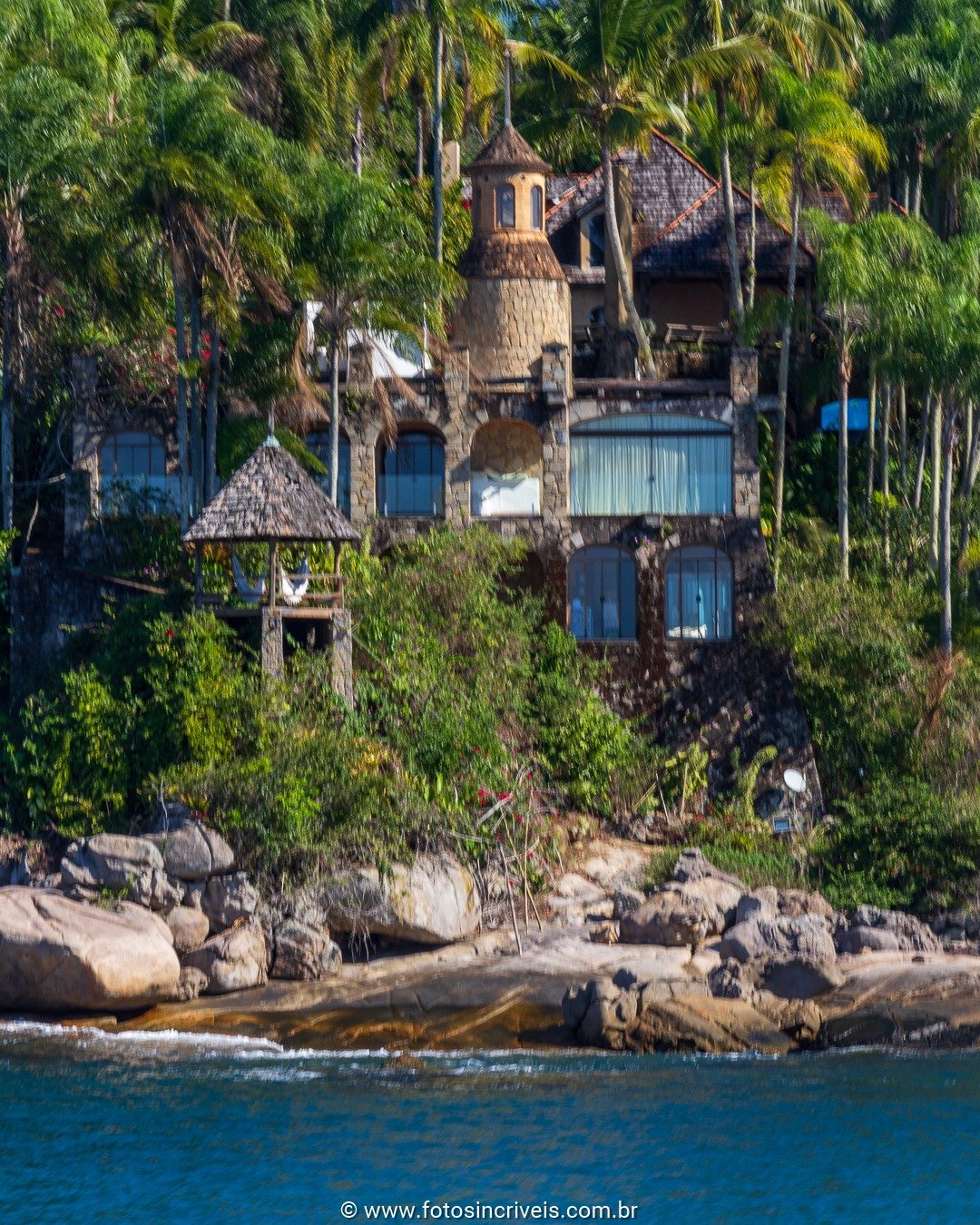 Casa de pedra com torre, construída sobre rochas à beira-mar, cercada por palmeiras e vegetação tropical densa.