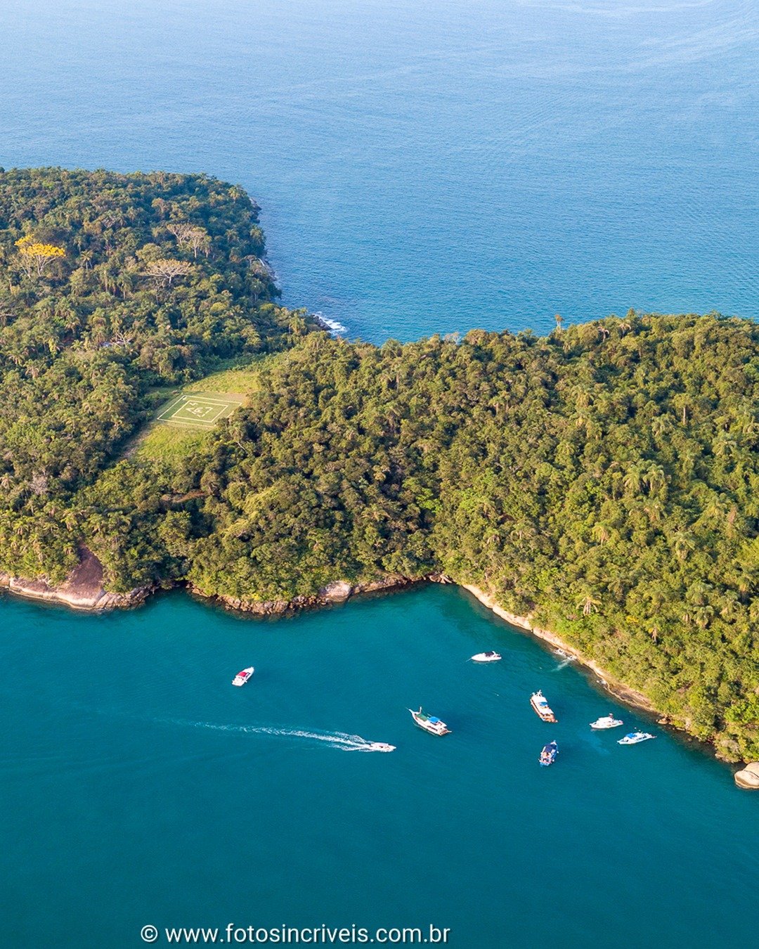 Vista aérea de uma ilha ou península coberta por floresta tropical, cercada por mar azul, com diversas embarcações ancoradas ao redor.