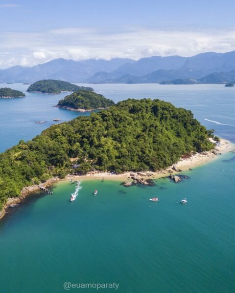 Vista aérea de ilha com vegetação densa, pequenas praias, água turquesa e embarcações, com montanhas ao fundo.