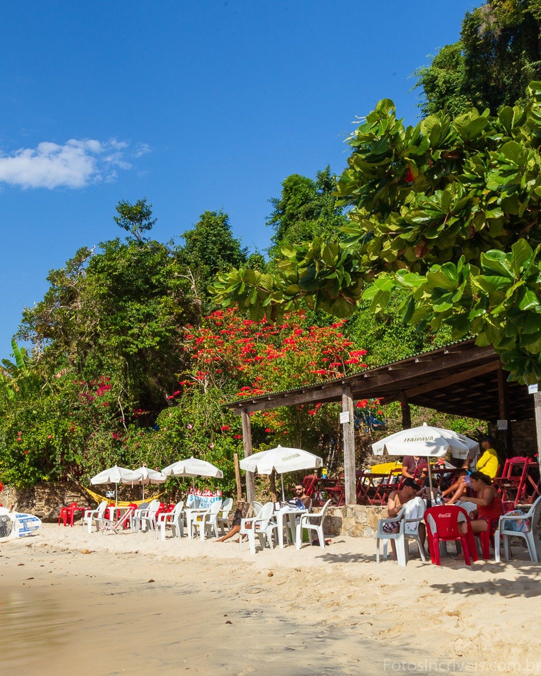 Praia ensolarada com mar calmo, barco amarelo na água, mesas e guarda-sóis na areia, quiosque rústico à direita e vegetação exuberante ao fundo.