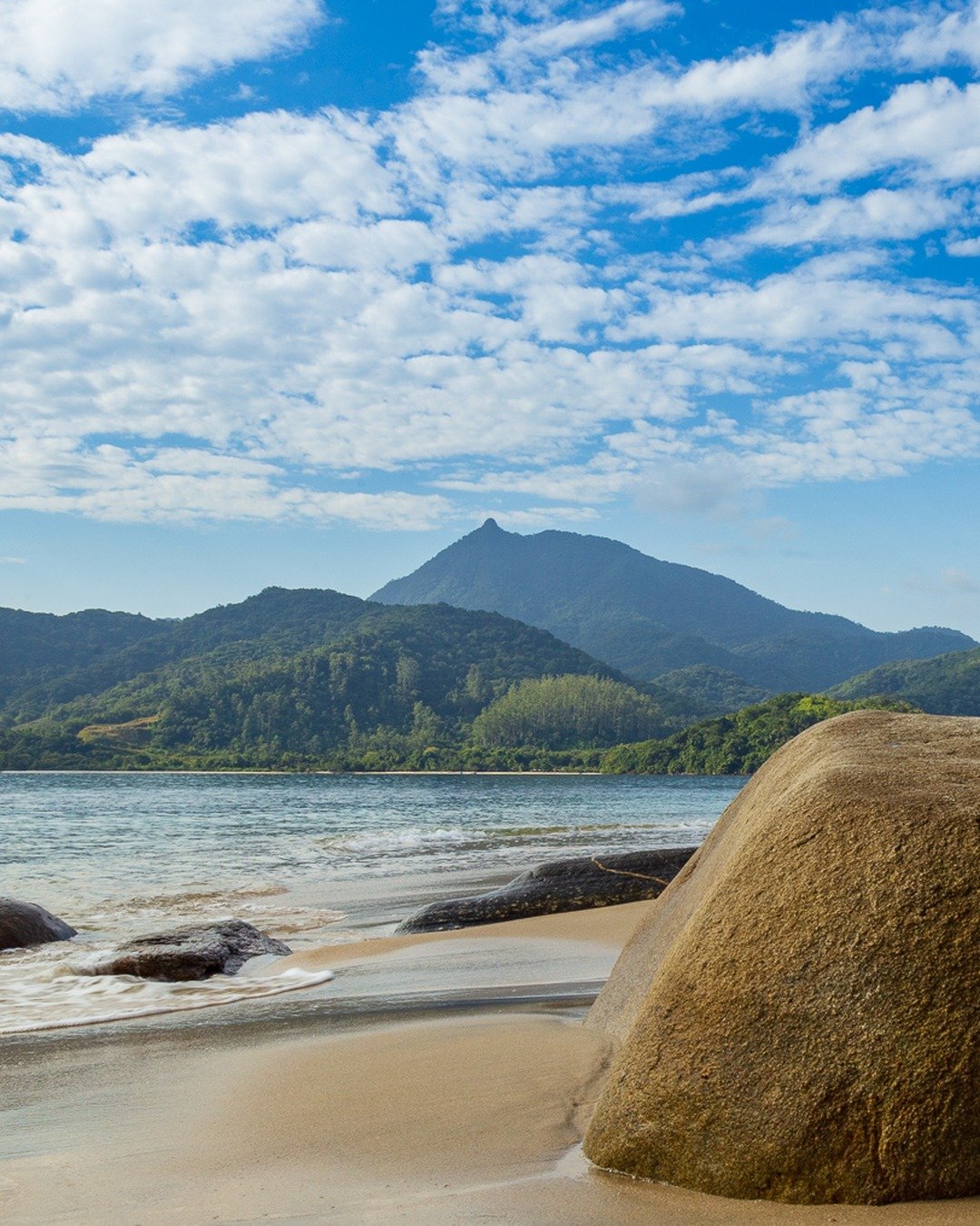 Praia de areia clara com grandes pedras arredondadas em primeiro plano, mar calmo e montanhas verdes ao fundo sob céu azul com nuvens esparsas.