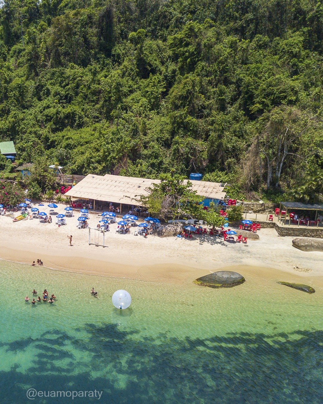 Vista aérea de uma praia de areia clara cercada por morro de mata atlântica, com quiosque de telhado rústico, guarda-sóis azuis e vermelhos, banhistas na água esverdeada e uma embarcação ancorada à esquerda.