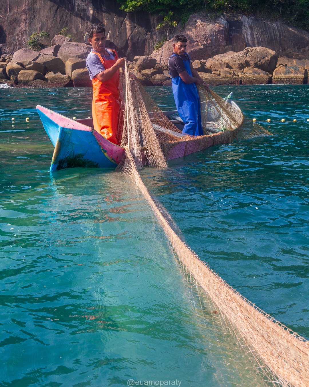 Dois pescadores em pequenos barcos coloridos puxam uma rede de pesca nas águas claras e turquesas junto a uma costa rochosa com vegetação densa ao fundo.