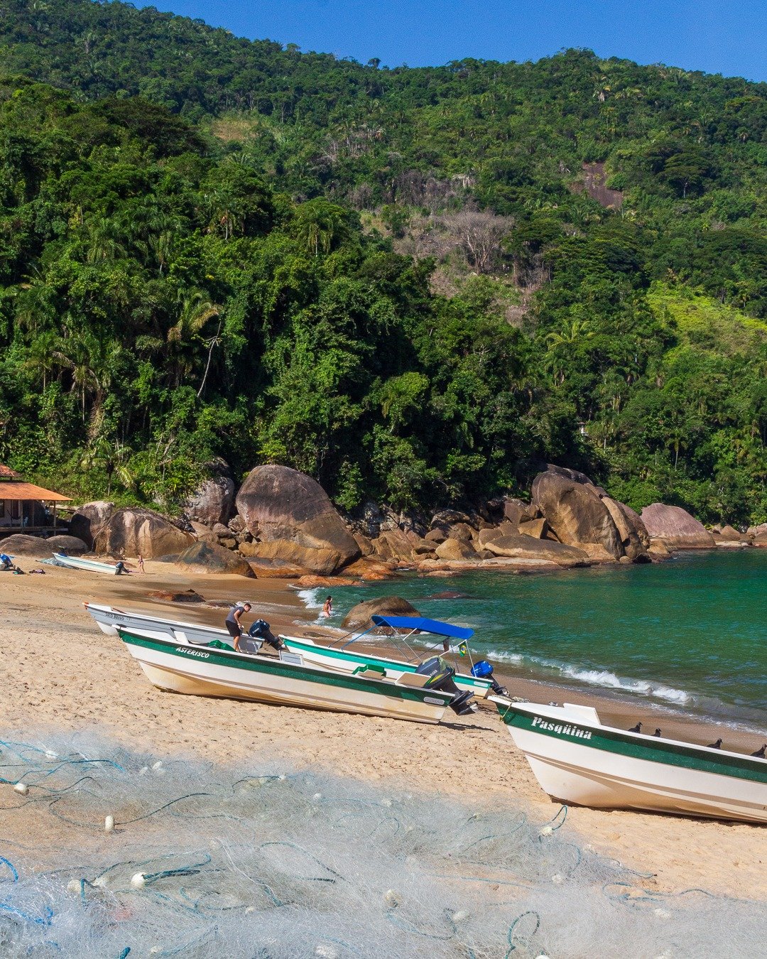Praia com barcos de pesca na areia, redes de pesca em primeiro plano e encosta de mata atlântica ao fundo.