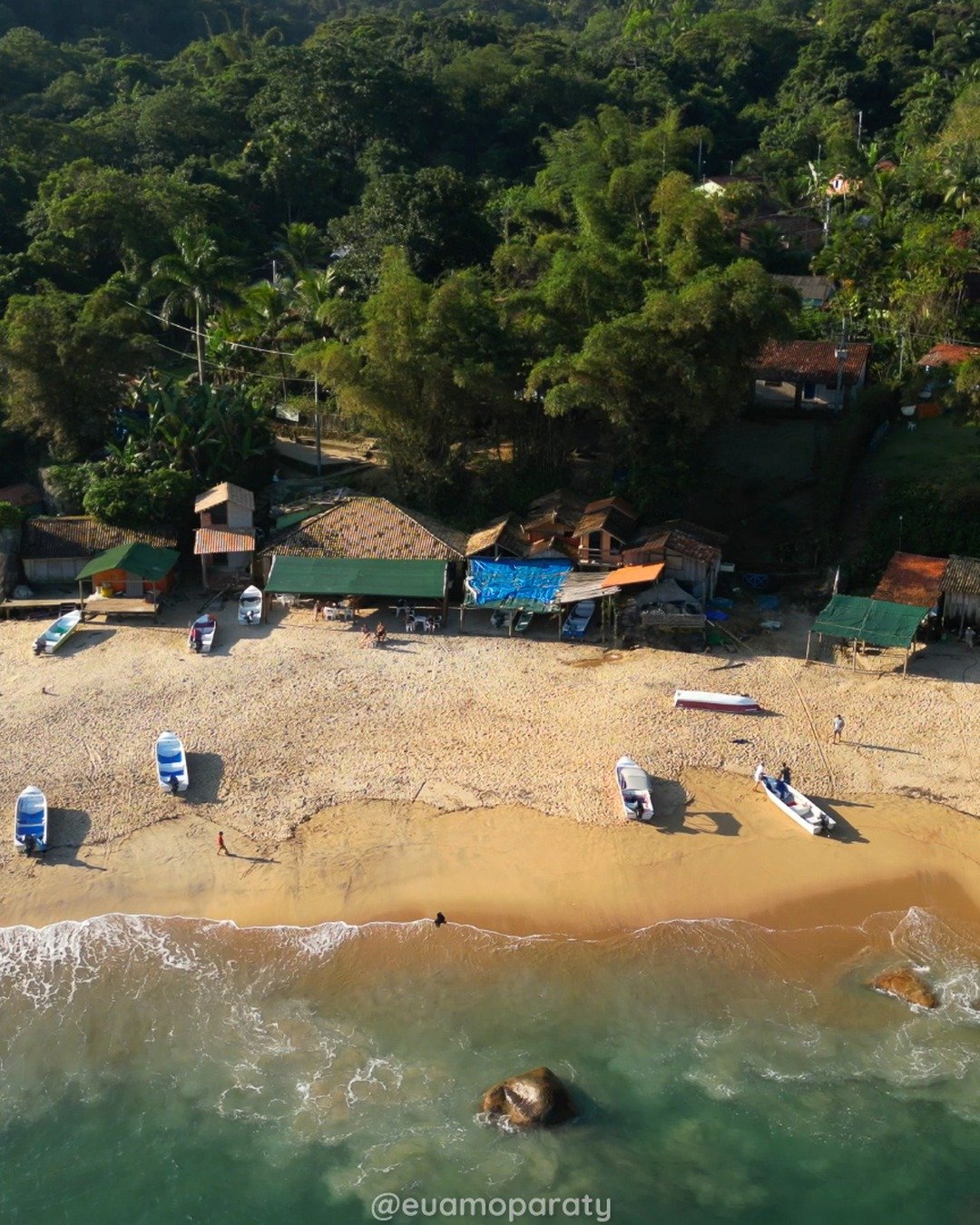 Vista aérea de uma praia com barcos, construções rústicas à beira-mar e mata densa ao fundo.