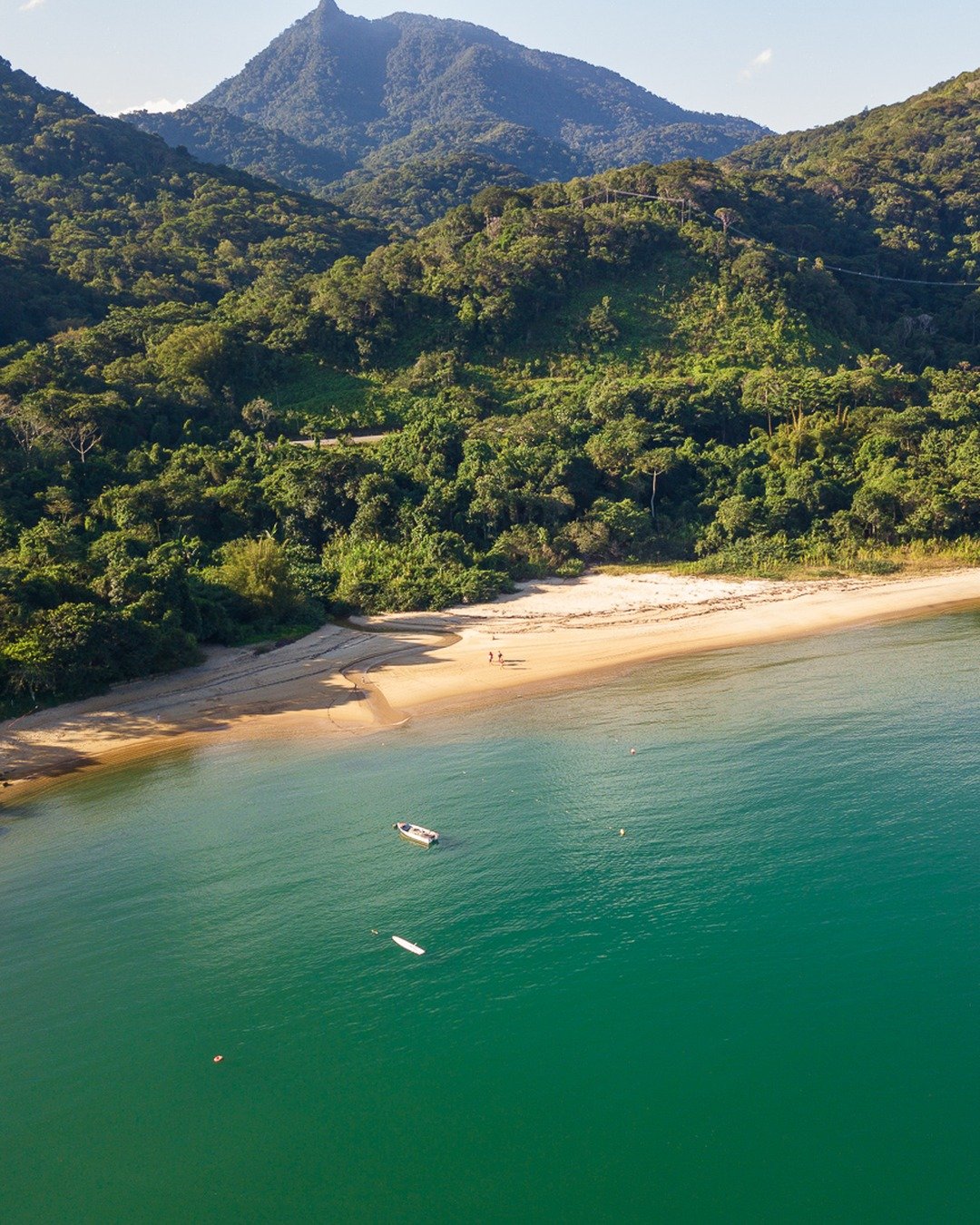 Vista aérea de uma enseada com praia de areia clara, mar verde-azulado e montanhas cobertas por mata atlântica.