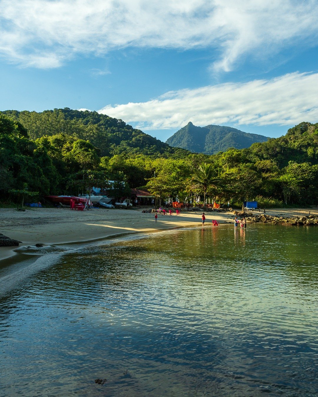 Praia de águas calmas com pessoas na areia e montanhas verdes cobertas por floresta ao fundo, sob céu azul com nuvens.