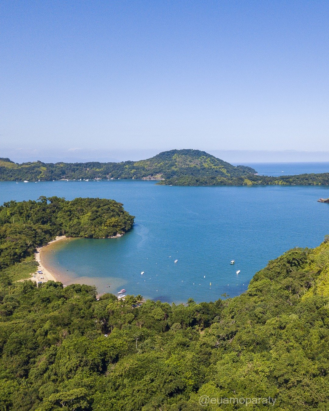 Vista elevada de uma baía costeira com ilhas verdes, pequena praia e barcos, sob céu azul.