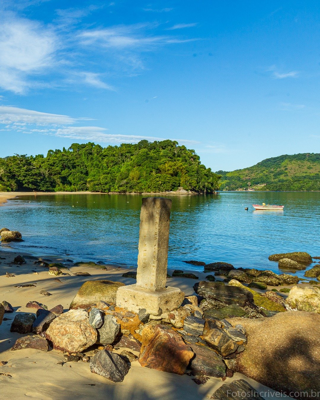 Praia de areia com rochas em primeiro plano, água calma, morros verdes ao fundo e um pequeno barco à direita.