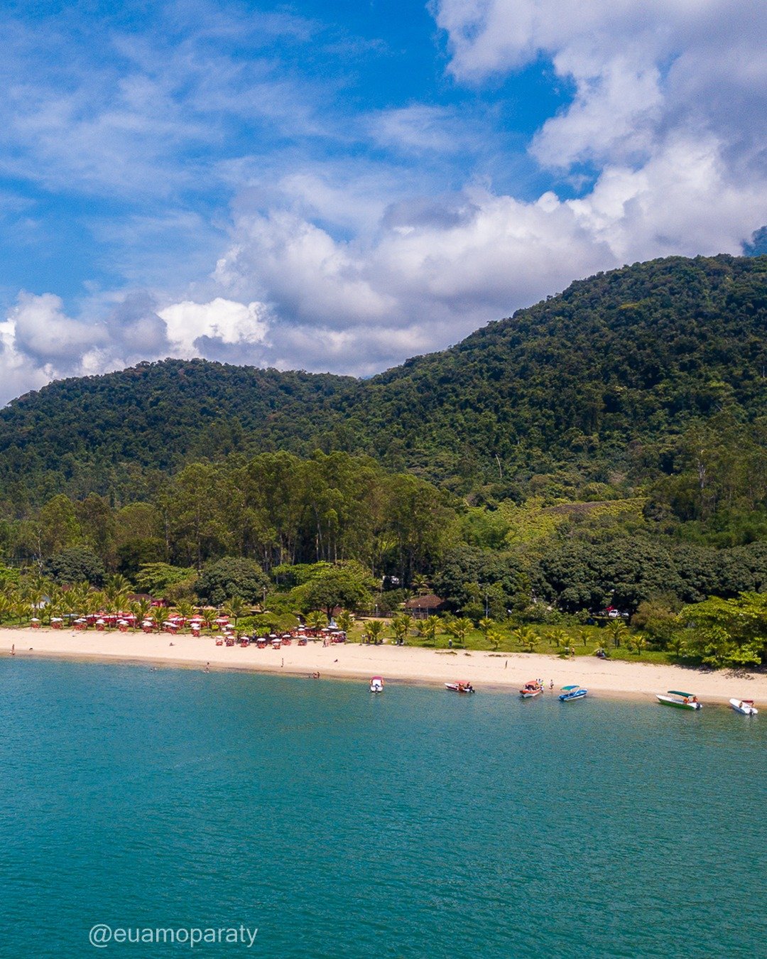 Vista panorâmica de uma enseada com praia de areia clara, mar turquesa, vegetação densa e montanhas ao fundo sob céu com nuvens.