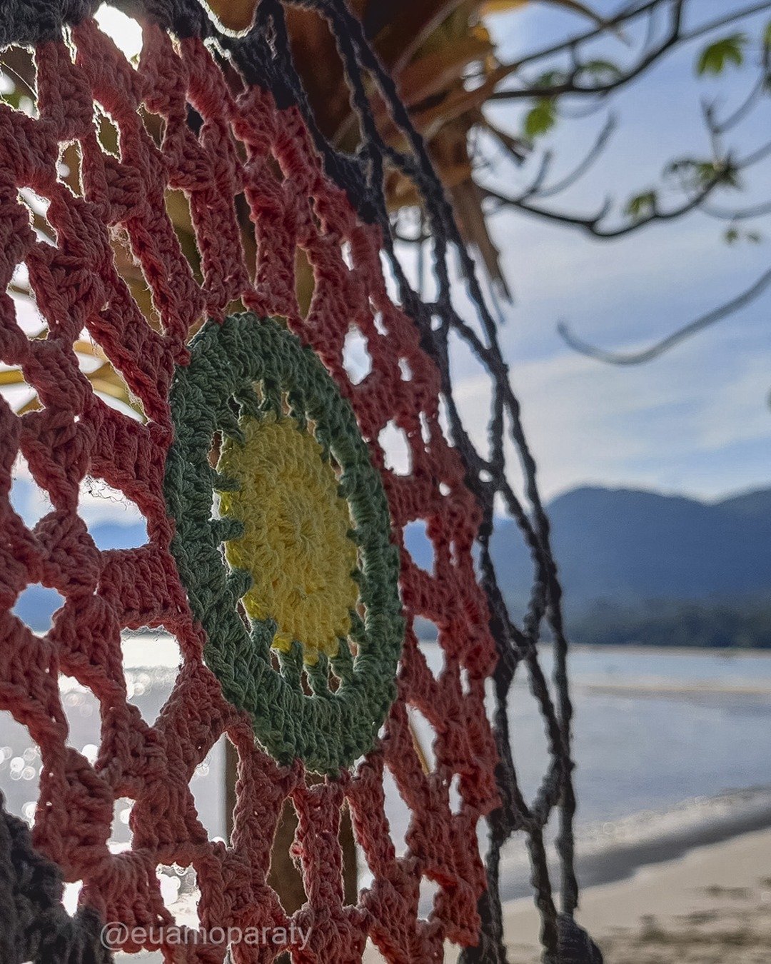 Arte em crochê colorida em primeiro plano, com praia, mar, montanhas e céu ao fundo.
