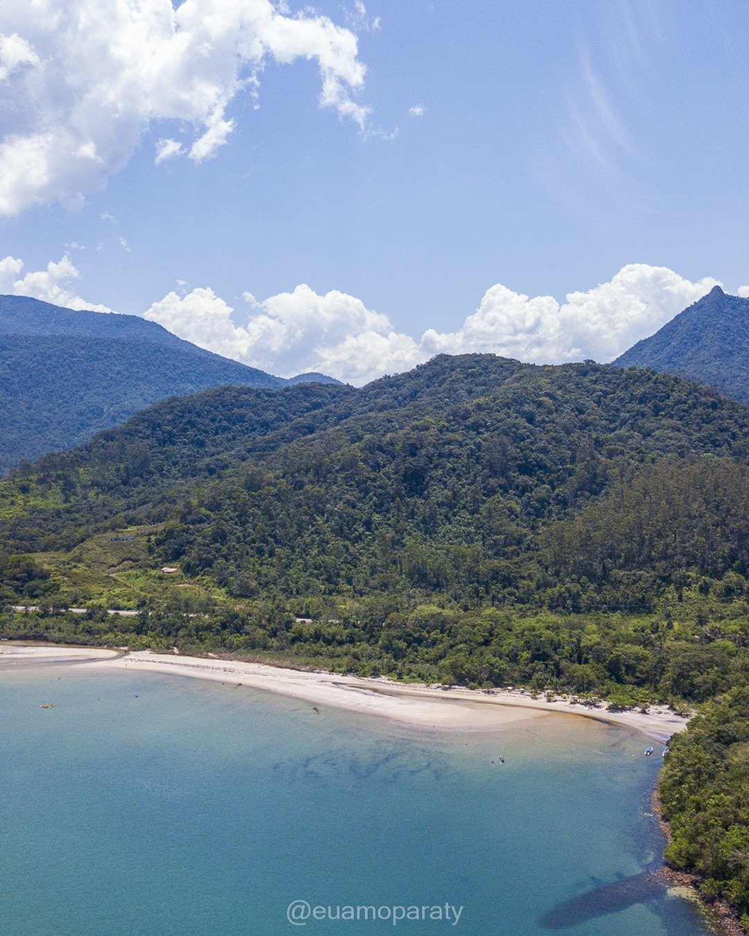 Vista aérea de uma enseada com praia de areia clara, mar azul-esverdeado e montanhas cobertas de Mata Atlântica ao fundo, sob céu com nuvens.