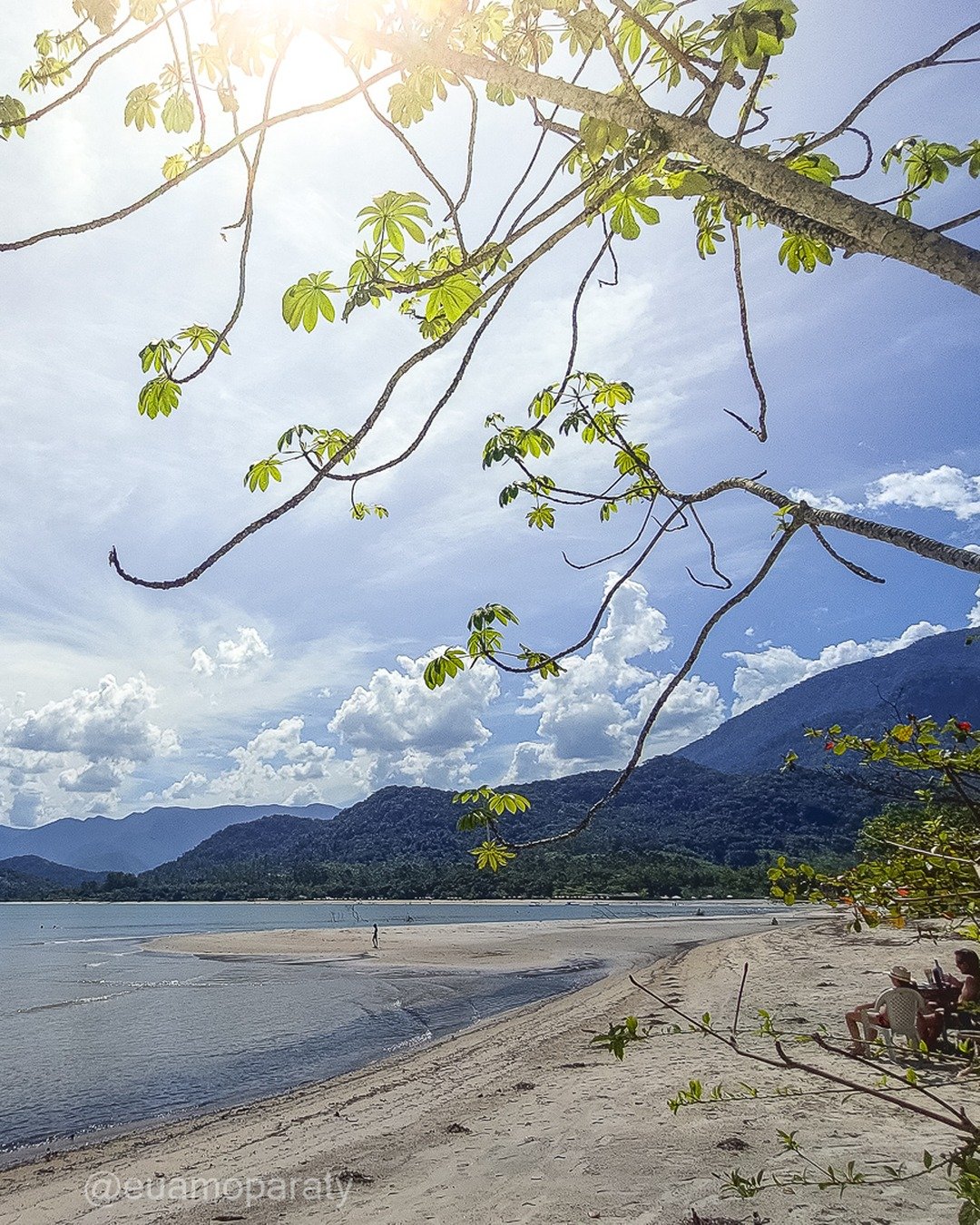 Praia com mar calmo, montanhas ao fundo, céu azul com nuvens e galhos de árvore em primeiro plano.