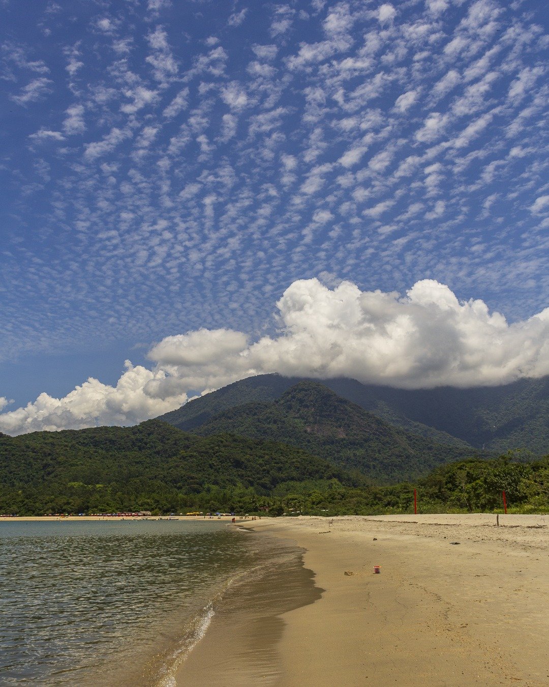 Praia com areia clara em primeiro plano, mar calmo à esquerda, montanhas cobertas de floresta ao fundo e céu azul com nuvens marcantes.