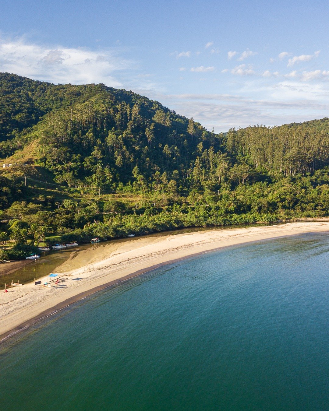 Vista aérea de uma enseada com praia de areia clara, mar calmo esverdeado e morros cobertos por vegetação ao fundo.