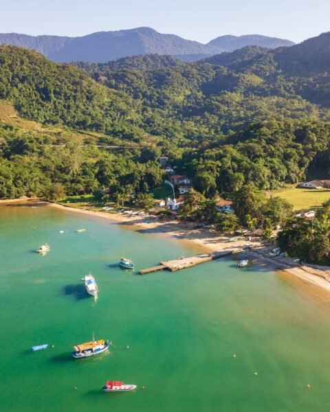 Vista aérea de uma enseada de águas verde-turquesa com barcos ancorados, faixa de areia, pequeno píer e casas cercadas por mata densa e montanhas ao fundo.