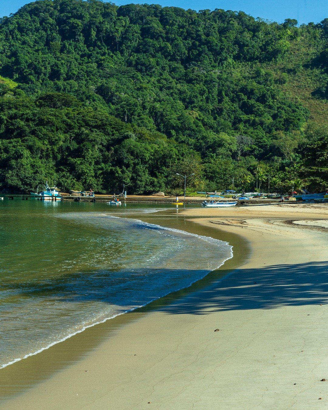 Praia de areia clara com mar calmo em tom esverdeado, faixa de sombra projetada na areia em primeiro plano, pequenas embarcações ancoradas próximas à margem e encosta de mata atlântica densa ao fundo sob céu azul limpo.