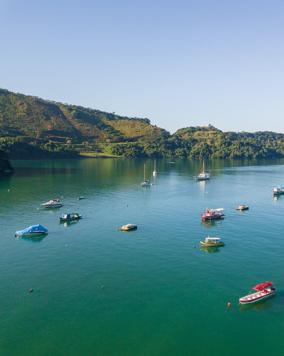Vista aérea de uma enseada de águas verde-escuras com diversas embarcações ancoradas, cercada por morros cobertos de vegetação sob céu azul claro.