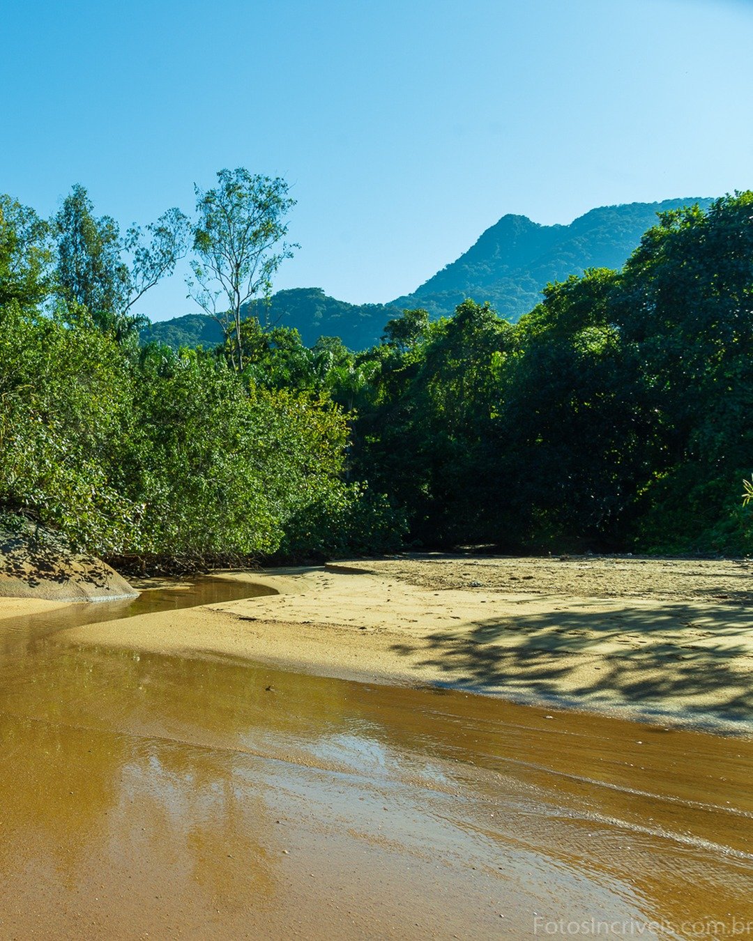Rio de águas douradas e calmas contornando bancos de areia, com grandes pedras arredondadas à esquerda, mata densa ao fundo e montanhas sob céu azul limpo à direita.