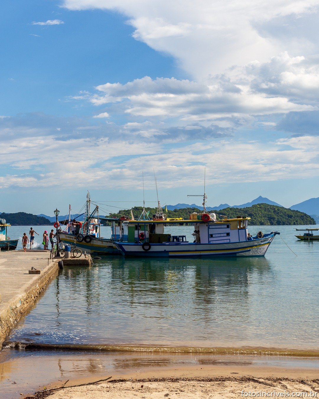 Píer de concreto com postes de iluminação à beira-mar, barcos ancorados e pessoas ao fundo, com morros verdes e céu azul com nuvens.