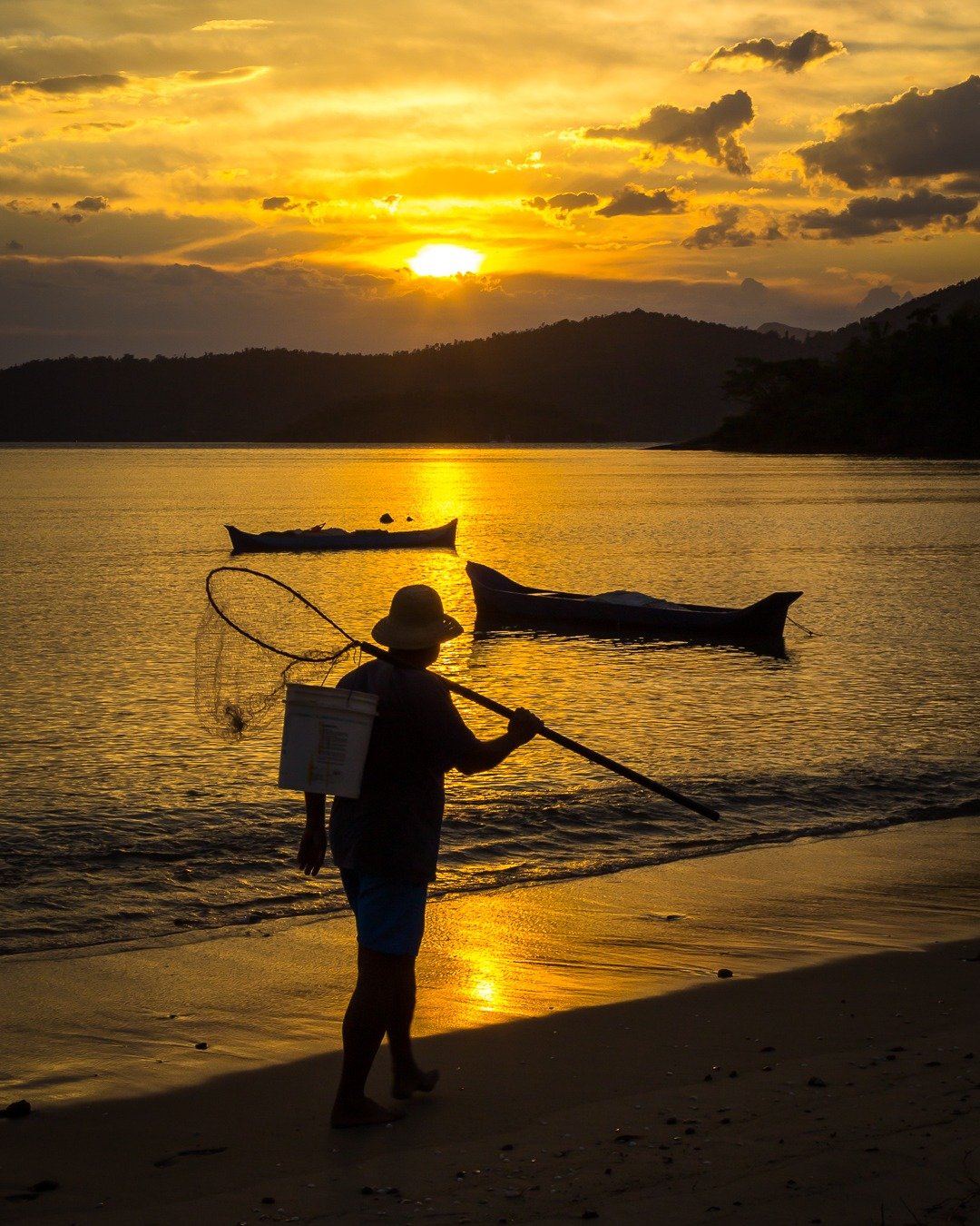 Silhueta de um pescador com rede de arrasto e caixa nas costas, caminhando na areia à beira-mar ao pôr do sol, com barcos ancorados na água dourada e montanhas ao fundo.