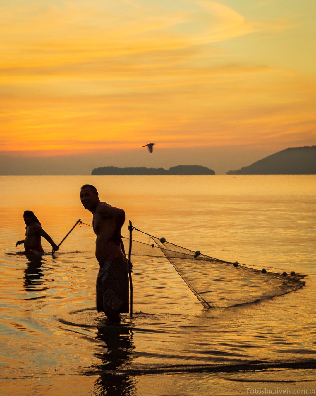 Dois pescadores em silhueta manuseando uma rede dentro da água durante o pôr do sol, com uma ave em voo e ilhas ao fundo.