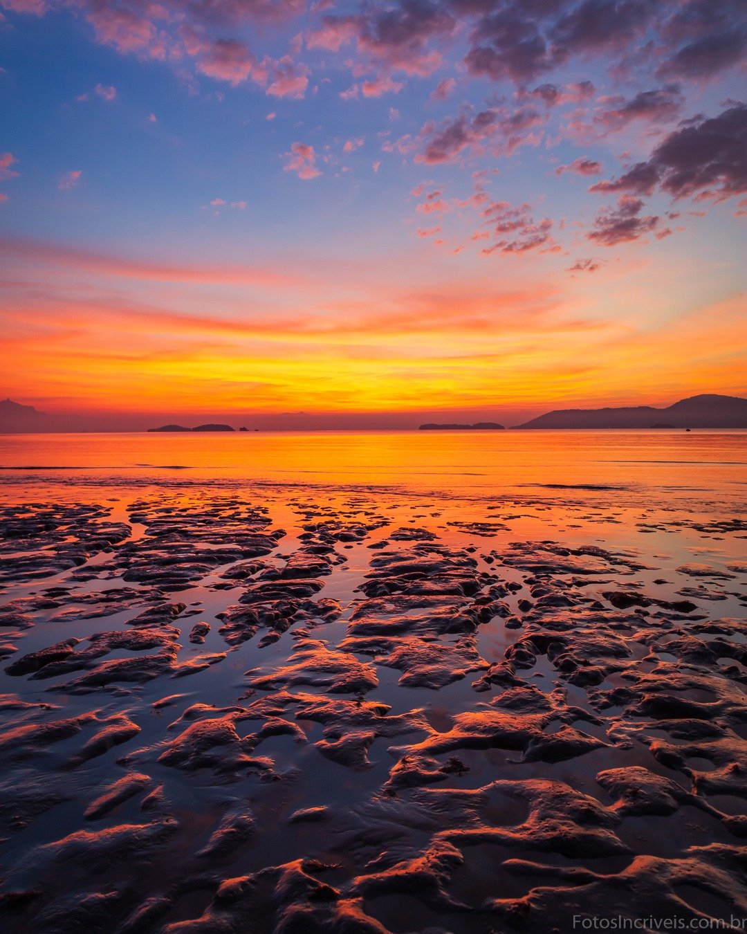 Sunset over calm water with a vibrant orange and purple sky, distant hills, and textured rippled sand/mudflats in the foreground.