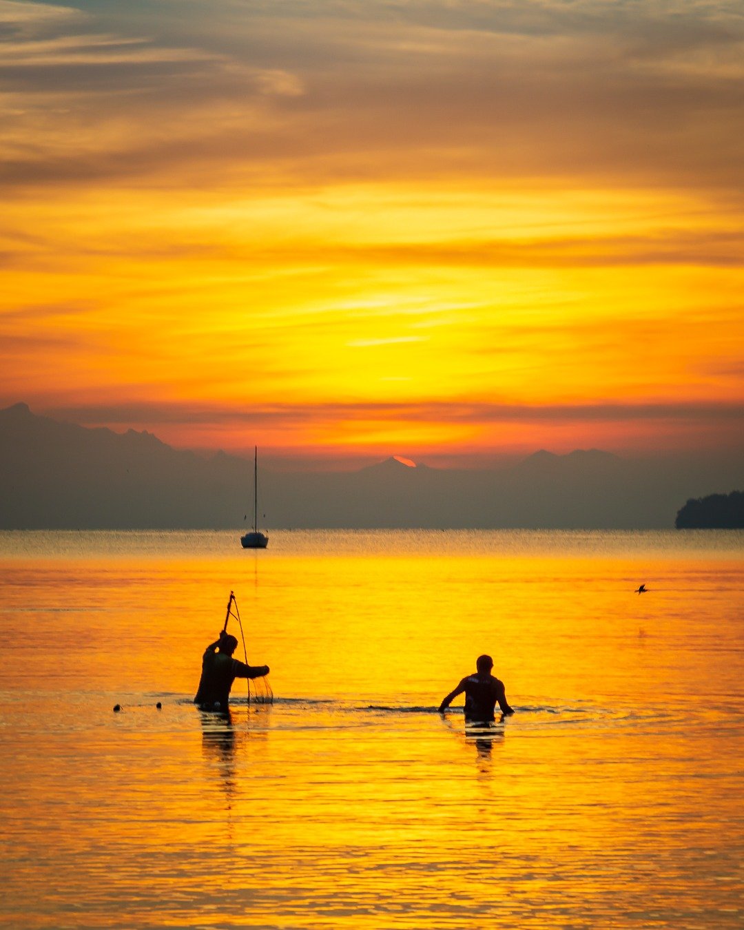 Pôr do sol intensamente dourado sobre o mar, com duas silhuetas humanas dentro da água rasa, um barco a vela ao fundo e montanhas no horizonte.