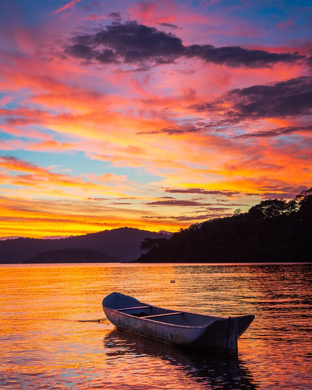 Canoa branca flutuando sobre água calma com reflexos alaranjados de um pôr do sol intenso, montanhas e vegetação em silhueta ao fundo.