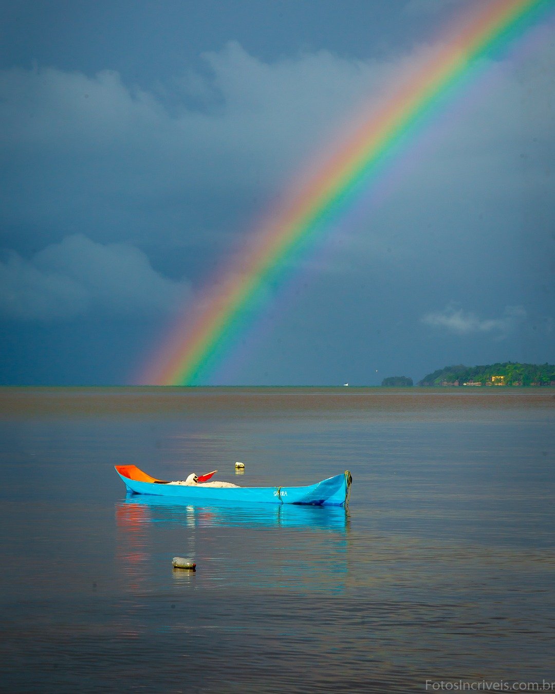 Pequeno barco azul flutuando sobre água calma e escura, sob céu carregado de nuvens com um arco-íris cruzando a diagonal da imagem; ao fundo, uma faixa fina de terra com vegetação no horizonte.