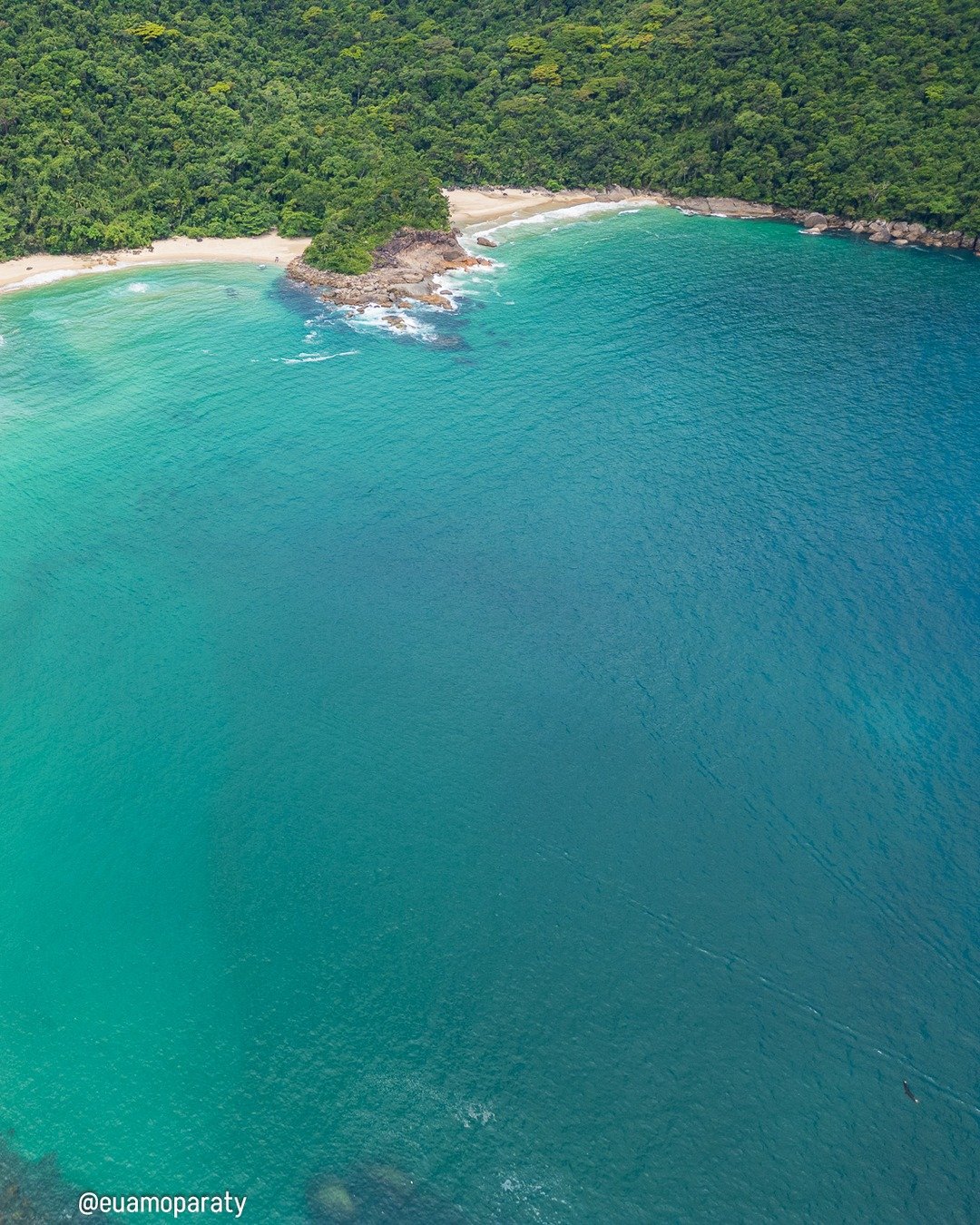 Vista aérea de uma enseada de águas turquesas com faixa de areia clara, rochas costeiras e floresta tropical densa contornando a praia.