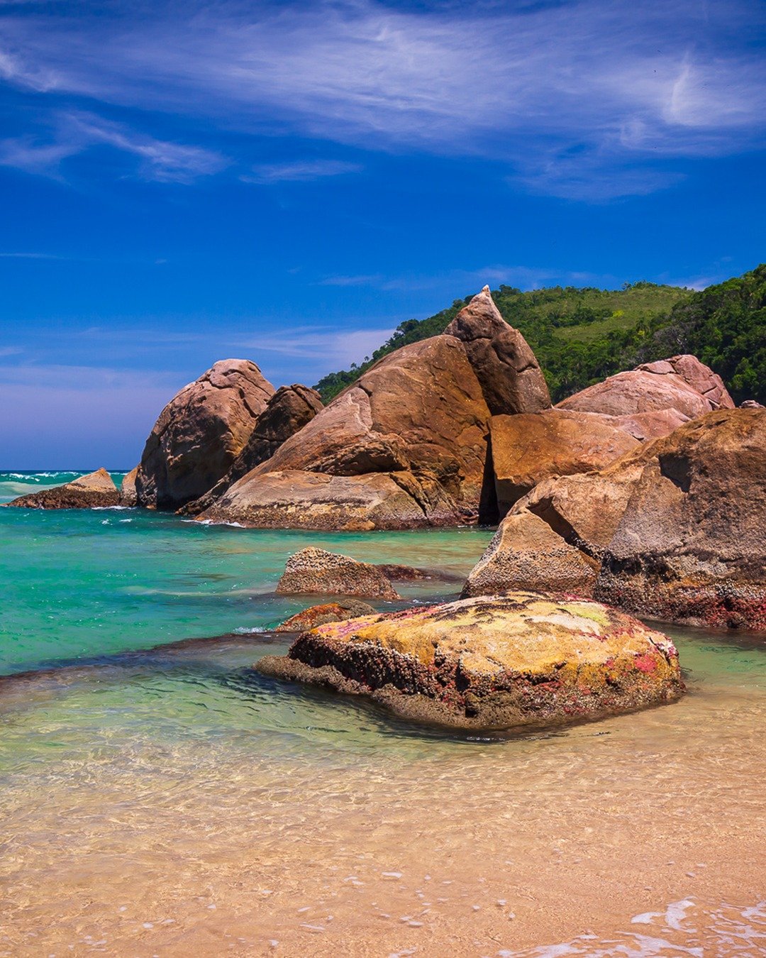 Praia com mar turquesa calmo, rochas graníticas avermelhadas em primeiro plano e encosta de mata verde ao fundo sob céu azul com nuvens.