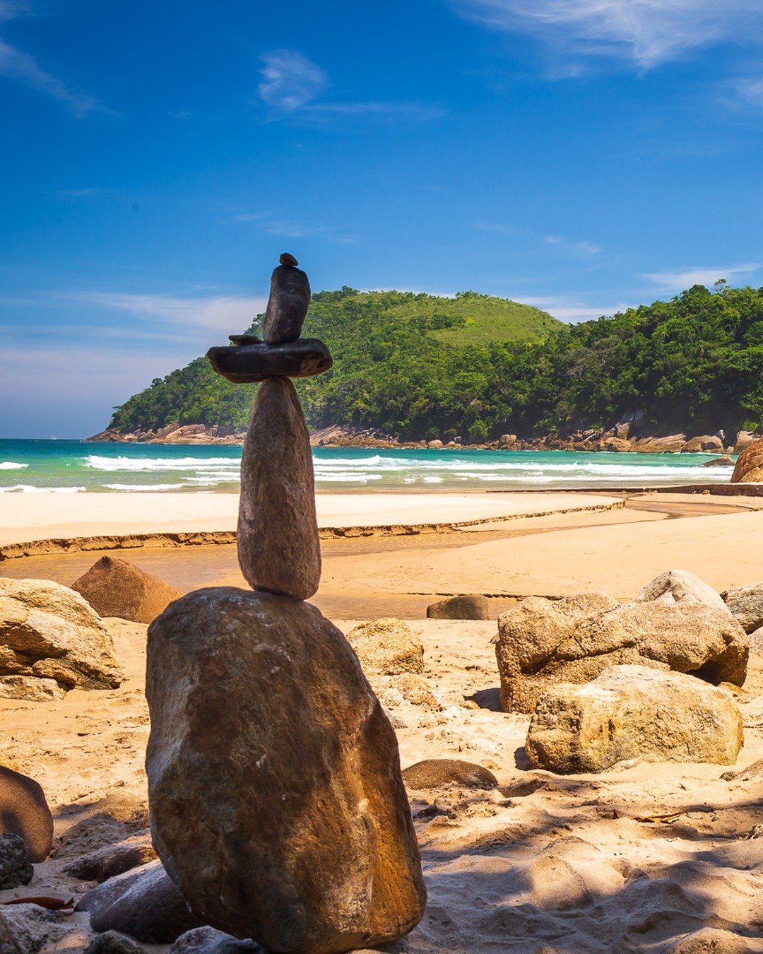 Praia de areia clara com pedras empilhadas em equilíbrio em primeiro plano, mar calmo com ondas ao fundo e morro coberto de vegetação densa sob céu azul com nuvens finas.