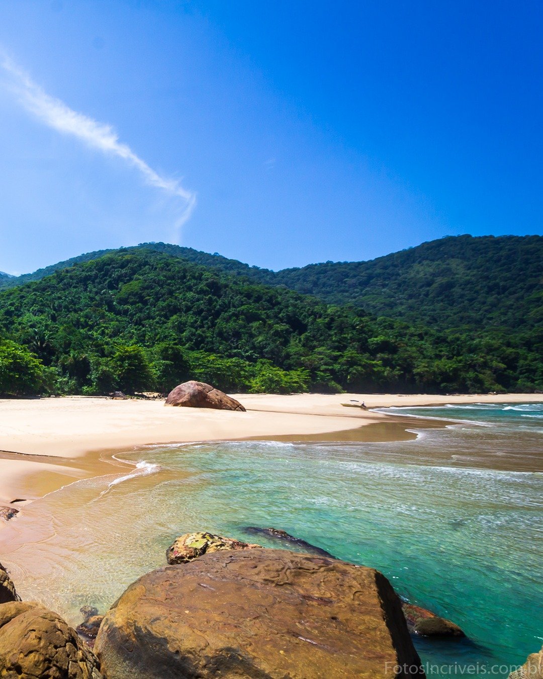 Praia de areia clara com rochas no primeiro plano, mar calmo e montanhas cobertas de mata ao fundo sob céu azul.