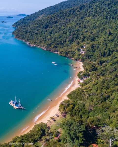 Vista aérea de uma enseada de água turquesa com faixa de areia contornando encosta de floresta densa; ilha arborizada ao fundo e embarcação ancorada próxima à praia.