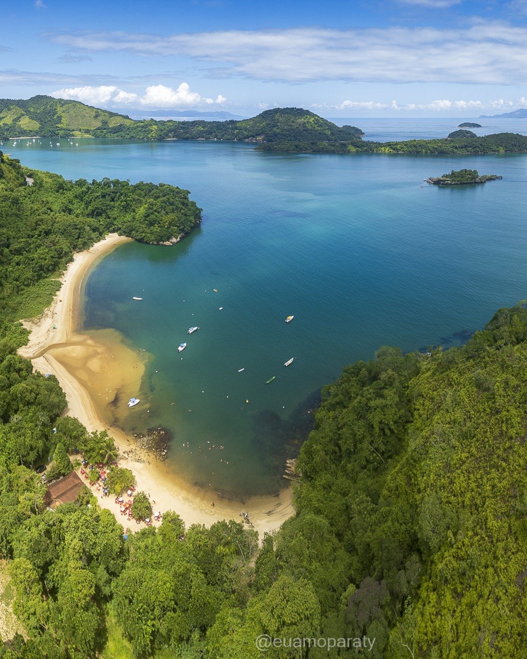 Vista aérea de uma enseada com praia de areia clara em curva, mar turquesa calmo com pequenas embarcações, cercada por morros cobertos de mata atlântica e ilhas ao fundo.