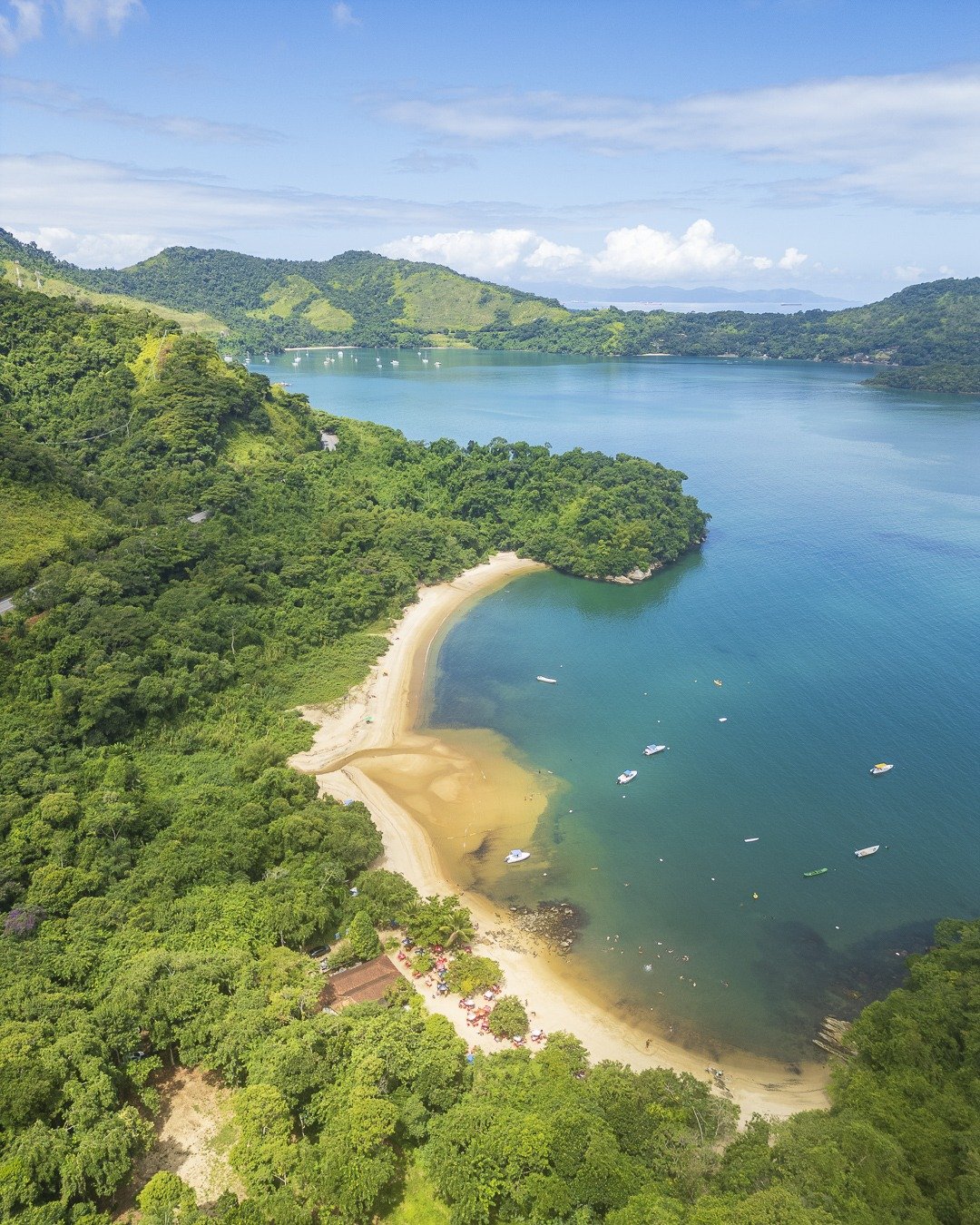 Vista aérea de enseada com praia de areia clara cercada por morros cobertos de vegetação densa, mar calmo esverdeado com pequenas embarcações e céu azul com nuvens.
