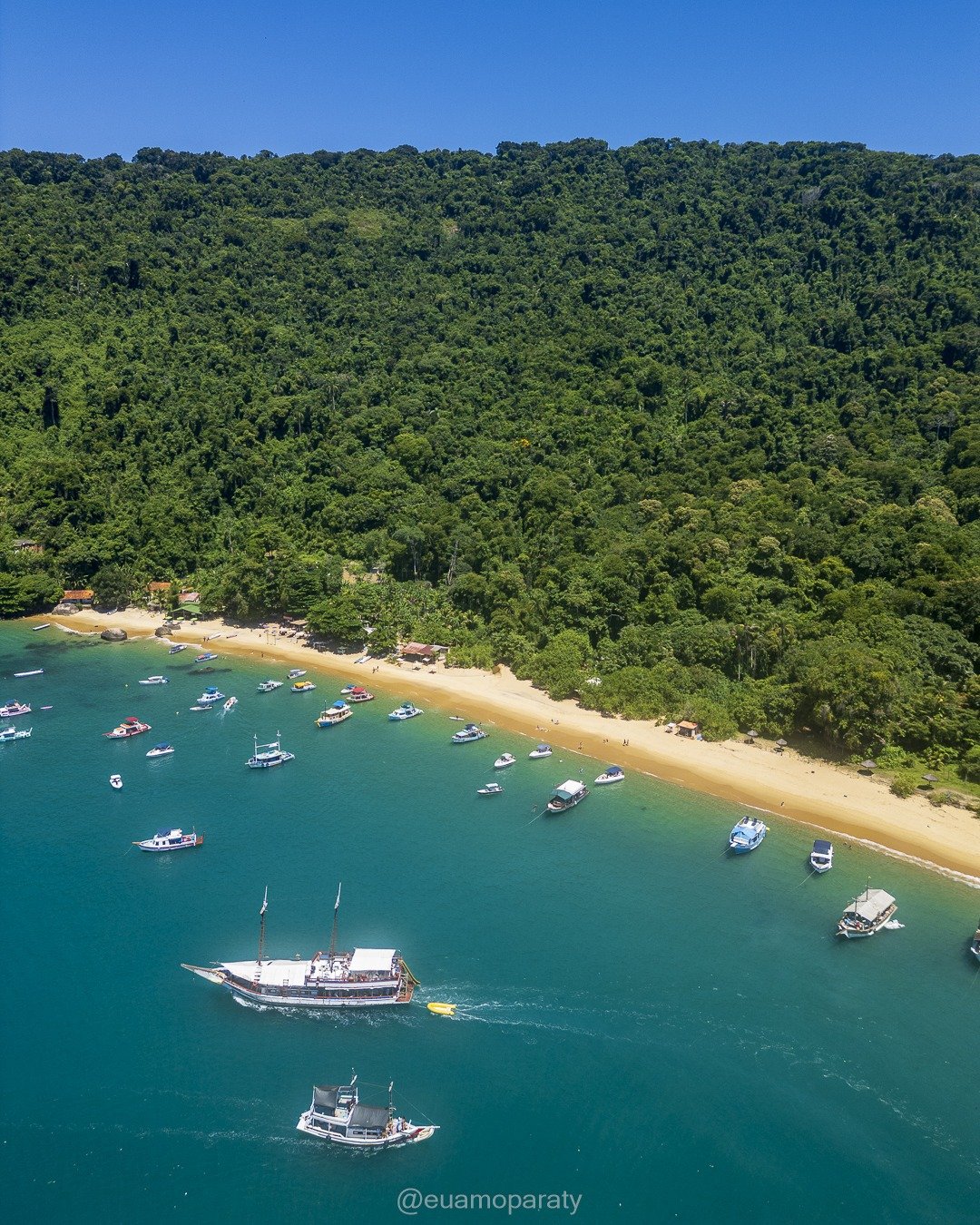 Vista aérea de uma enseada com faixa de areia à beira de mar turquesa repleto de embarcações ancoradas, tendo ao fundo uma encosta íngreme coberta por mata atlântica densa sob céu azul claro.