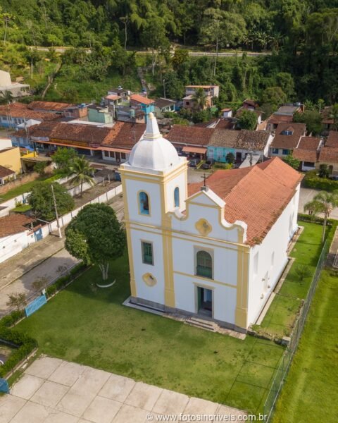 Vista aérea de uma igreja colonial branca com torre e cúpula, cercada por casario histórico e área verde, com morro florestado ao fundo.
