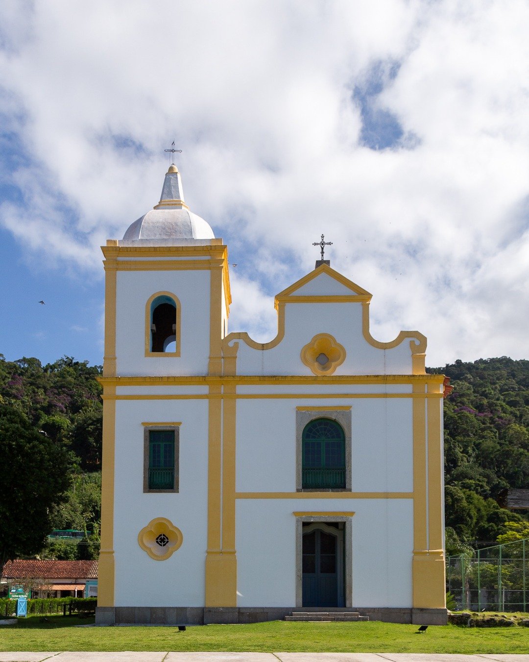 Igreja branca com detalhes amarelos e torre sineira à esquerda, sob céu azul com nuvens e morro verde ao fundo.