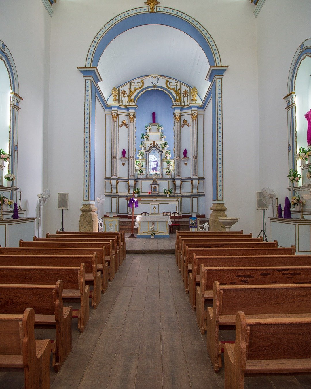 Interior de igreja católica com corredor central ladeado por fileiras de bancos de madeira, conduzindo ao altar-mor ornamentado em azul e branco, sob arco decorado e teto claro.