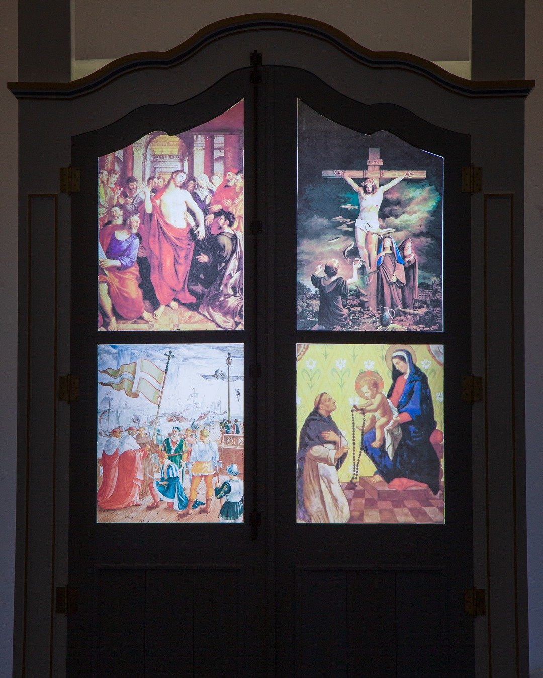 Peça de madeira escura com quatro painéis pintados representando cenas religiosas, vista em ambiente interno e pouco iluminado de igreja.