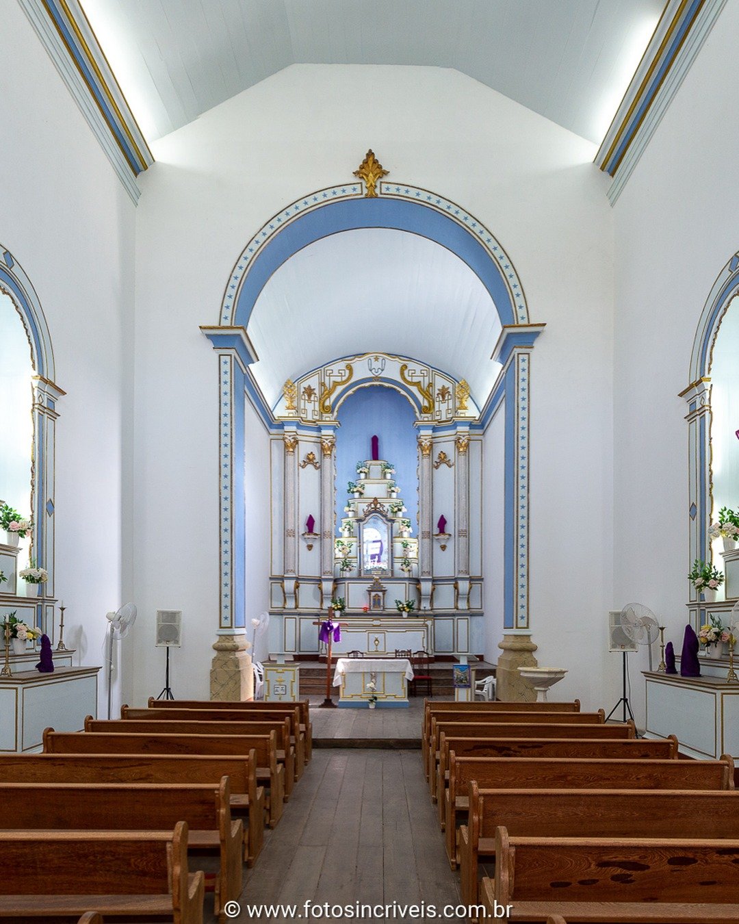 Interior de igreja de nave única, com paredes brancas e molduras azuis, fileiras de bancos de madeira e altar ao fundo sob arco azul e dourado.