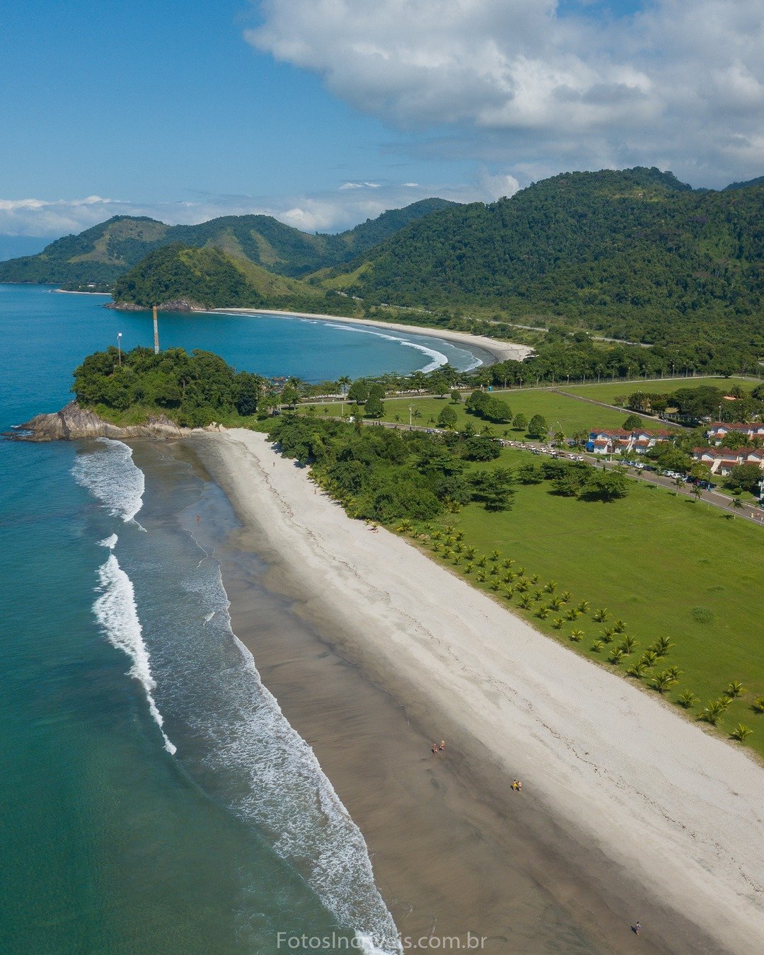 Vista aérea de uma praia com ondas quebrando na areia, faixa de campo verde, casas com telhados vermelhos, morros cobertos de vegetação e mar azul-turquesa sob céu com nuvens.