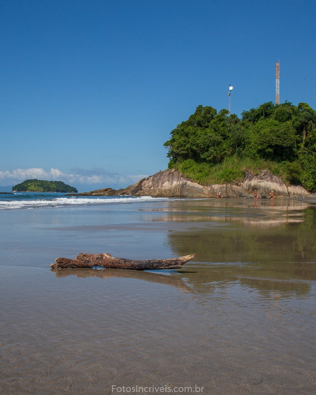Praia de areia molhada com reflexos do céu, tronco de madeira em primeiro plano e promontório rochoso coberto de vegetação à direita, com antena no topo; mar calmo e ilhas ao fundo.